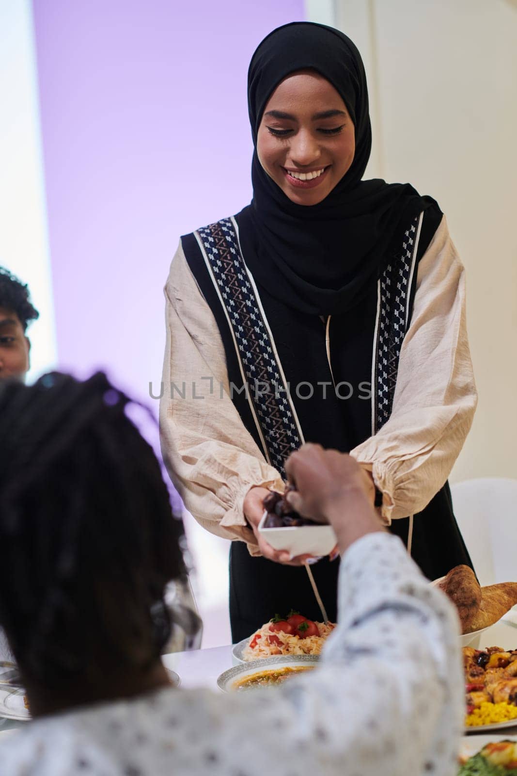 In a heartwarming scene during the sacred month of Ramadan, a traditional Muslim woman offers dates to her family gathered around the table, exemplifying the spirit of unity, generosity, and cultural richness during this festive and spiritual occasion.
