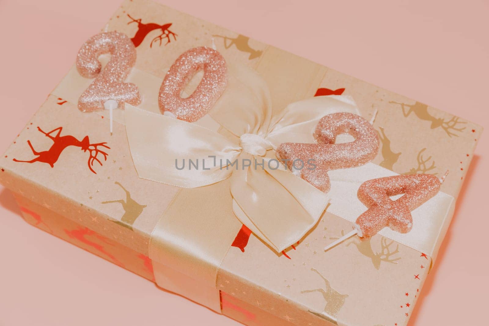 Shiny candles with the number 2024 on a gift box tied with a bow lie diagonally on a pink background, close-up side view.