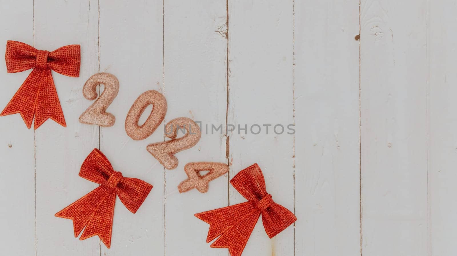 One festive light brown shiny candle number 2024 and three red Christmas bows lie on the left on a white wooden table with copy space on the right, flat lay close-up.