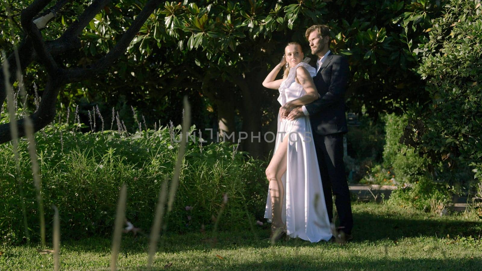 The bride and groom sitting by the tree trunk and hugging in the park. Action. Concept of romance and love, wedding traditions. by Mediawhalestock