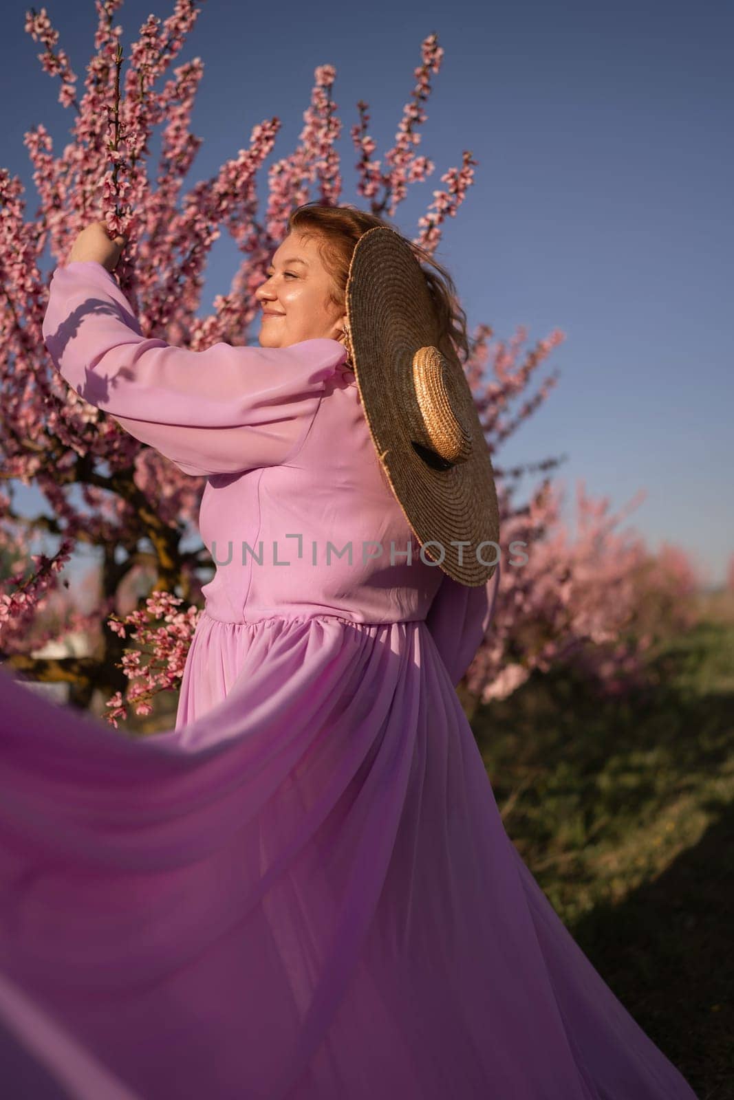 Woman blooming peach orchard. Against the backdrop of a picturesque peach orchard, a woman in a long pink dress and hat enjoys a peaceful walk in the park, surrounded by the beauty of nature. by Matiunina