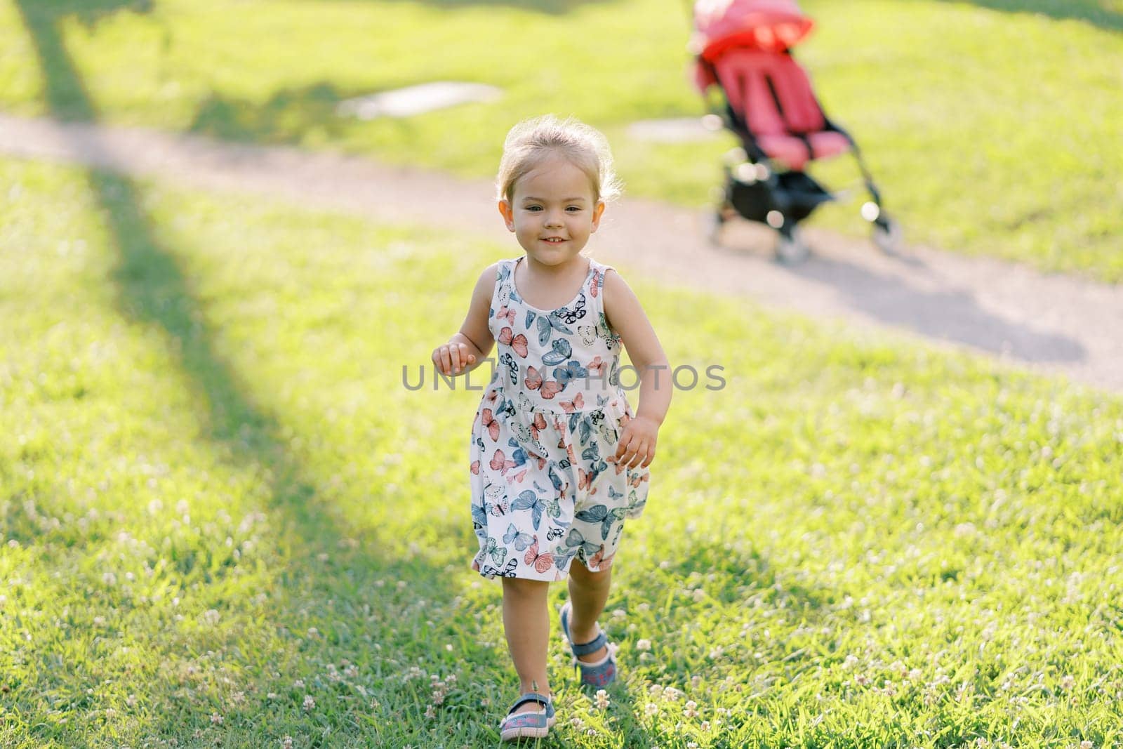 Little girl walks smiling along the green lawn from the stroller on the path by Nadtochiy