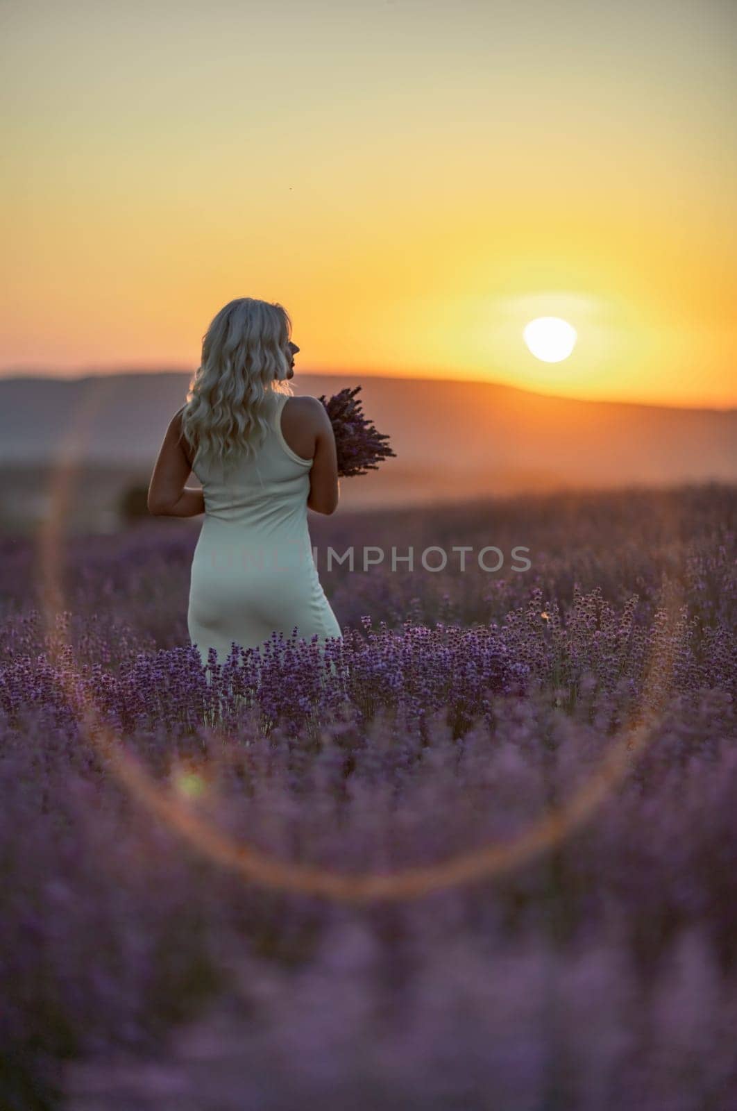 Blonde woman poses in lavender field at sunset. Happy woman in white dress holds lavender bouquet. Aromatherapy concept, lavender oil, photo session in lavender.