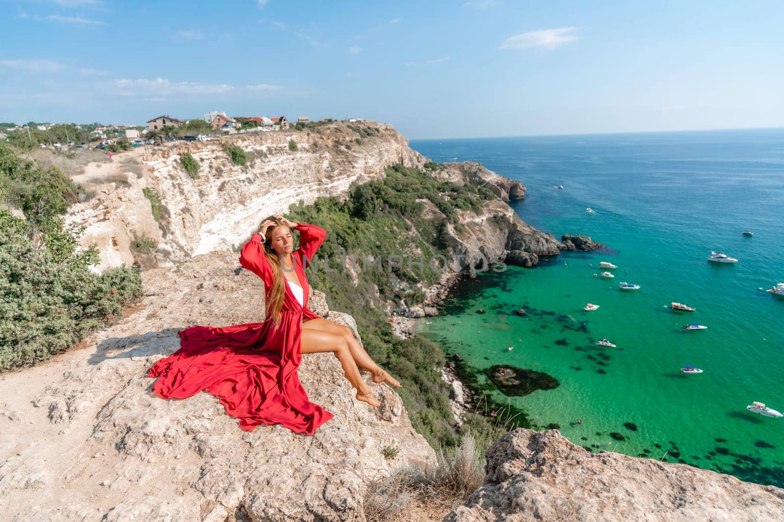 Woman red dress sea. Happy woman in a red dress and white bikini sitting on a rocky outcrop, gazing out at the sea with boats and yachts in the background