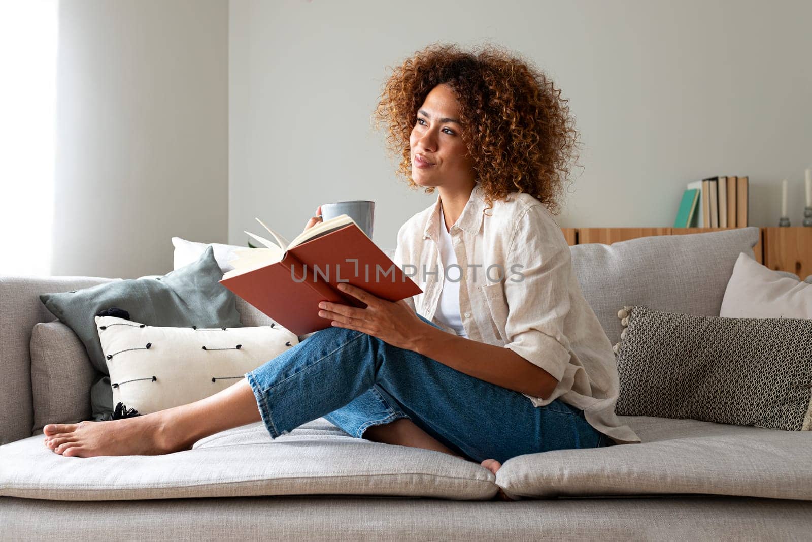 Pensive relaxed African american woman reading a book at home, drinking coffee sitting on the couch. Copy space. by Hoverstock