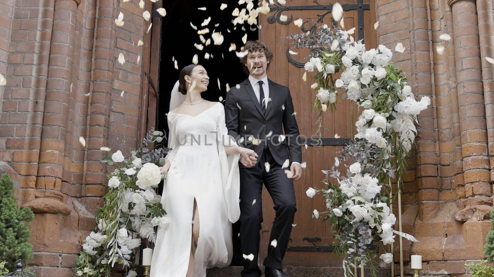 Just married young happy couple exiting church with the doors decorated by flowers. Action. Throwing white petals to man and woman. by Mediawhalestock