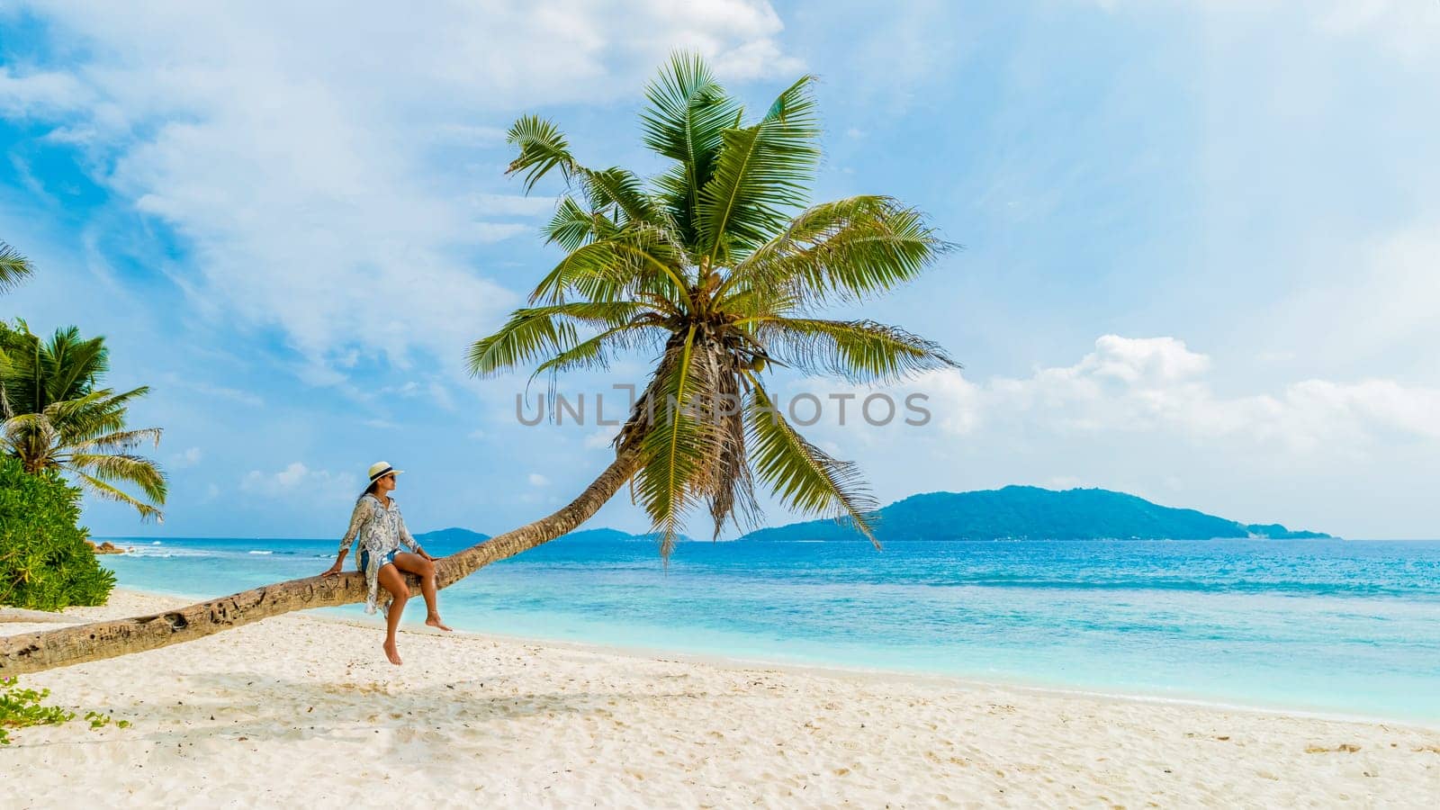 Young woman relaxing at a coconut palm tree on a white tropical beach at La Digue Seychelles Islands. Asian woman with hat sitting on a palm tree looking out over the ocean