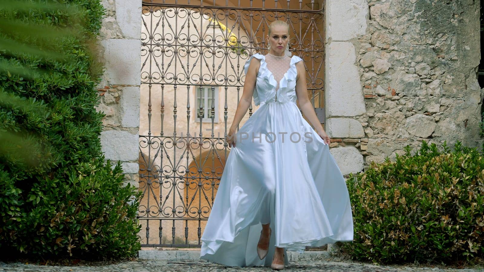 Bride walking in front of the gate in the city park. Action. Beautiful woman in white long dress near stone building. by Mediawhalestock