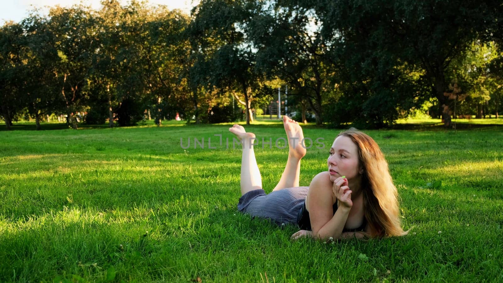 A cheerful woman in a clearing. Concept. A beautiful woman with long curly hair lying on the grass swinging her legs and laughing. by Mediawhalestock