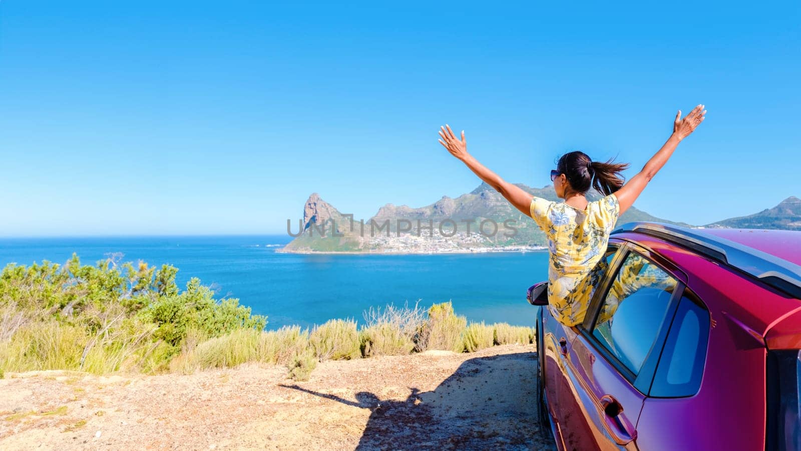 woman outside a car window with hands up, a car at Chapman's Peak Drive in Cape Town South Africa looking out over the ocean. women on a road trip at the garden route South Africa with a renal car