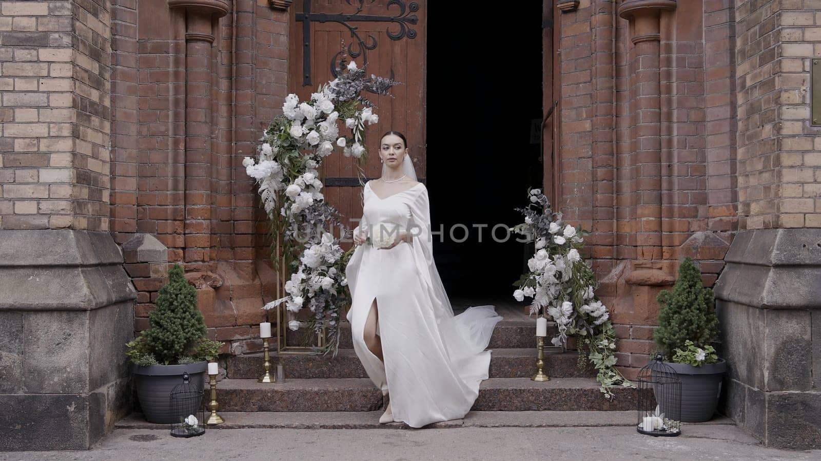 A young bride coming out of their temple. Action.A beautiful girl in a white dress with flowers who comes out of a dark building and there is an arch of white flowers nearby. by Mediawhalestock
