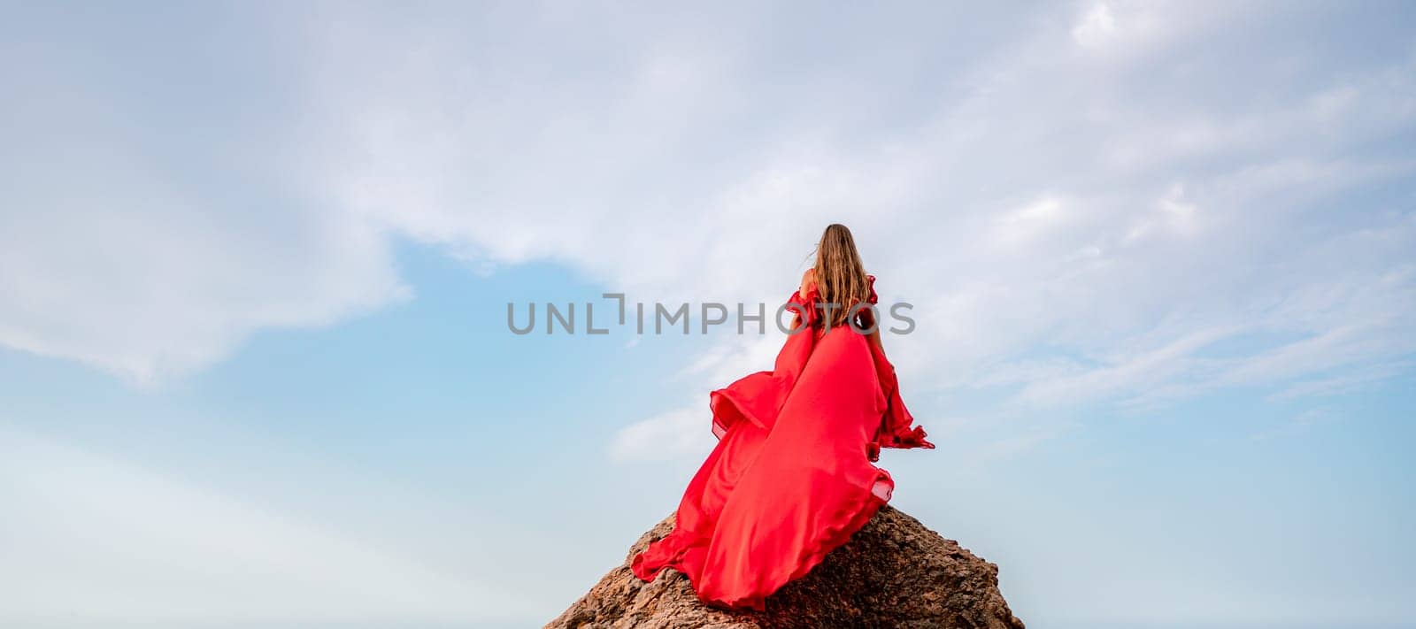 woman sky red dress. Woman with long hair on a sunny seashore in a red flowing dress, back view, silk fabric waving in the wind. Against the backdrop of the blue sky and mountains on the seashore. by Matiunina