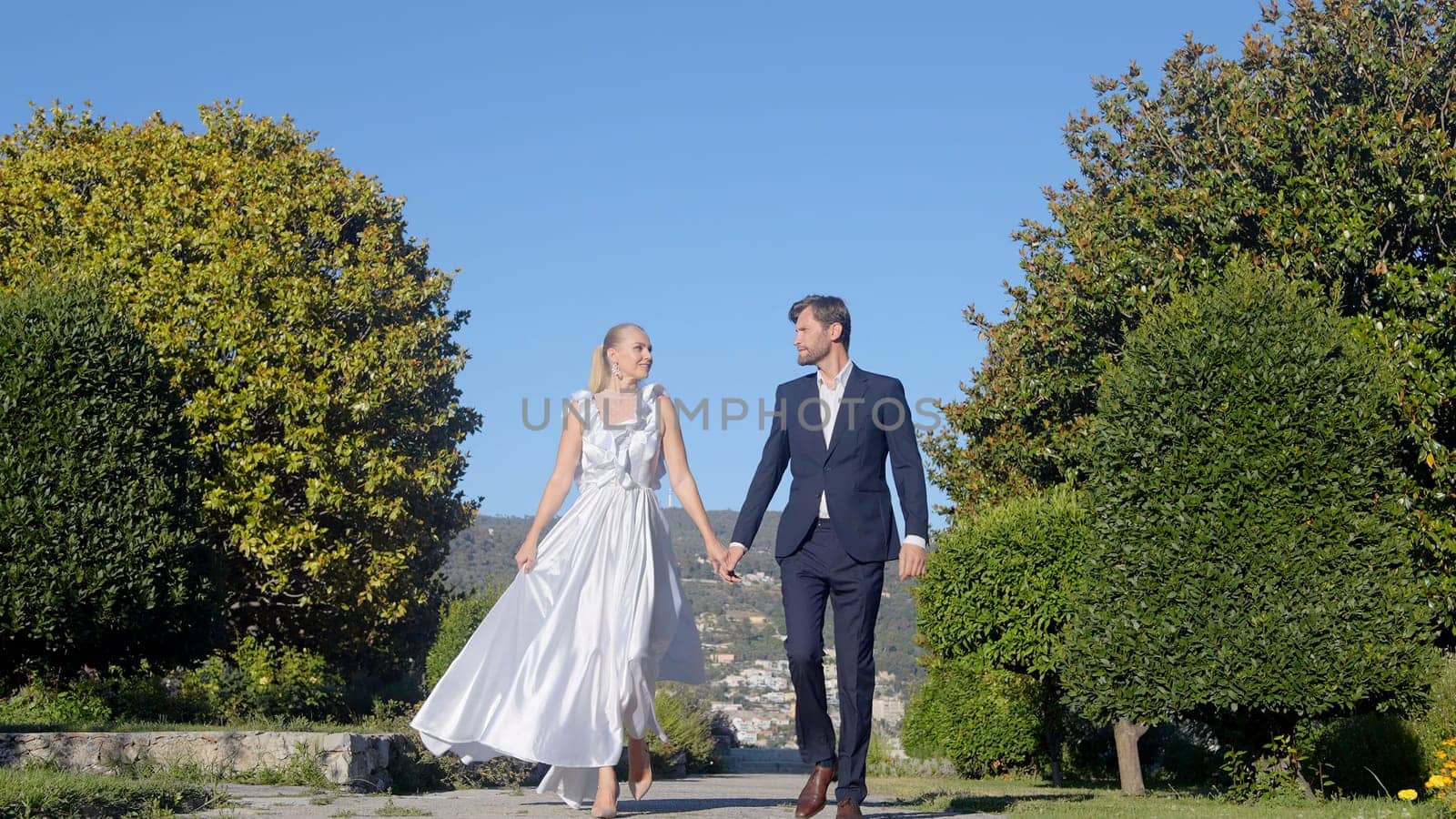 Happy bride and groom walking and holding hands in the park. Action. Wedding couple during video shooting among green summer vegetation. by Mediawhalestock