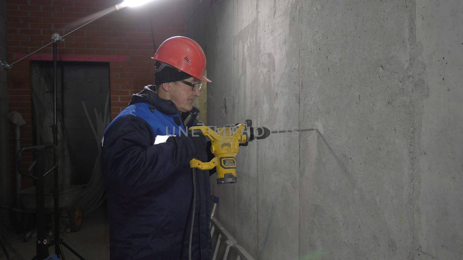Builder in uniform drills hole in wall. Clip. Repairman makes hole with drill. Builder with drill makes hole in new room.