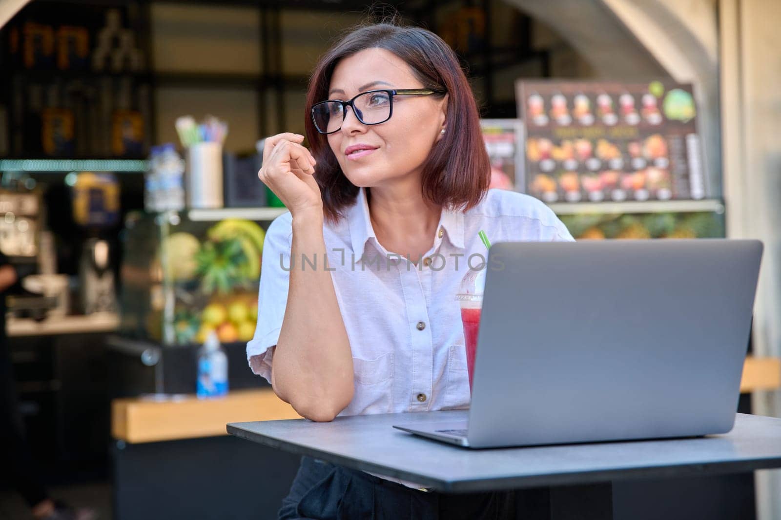 Middle-aged woman with laptop at table in an outdoor fruit juice bar with fresh juice by VH-studio