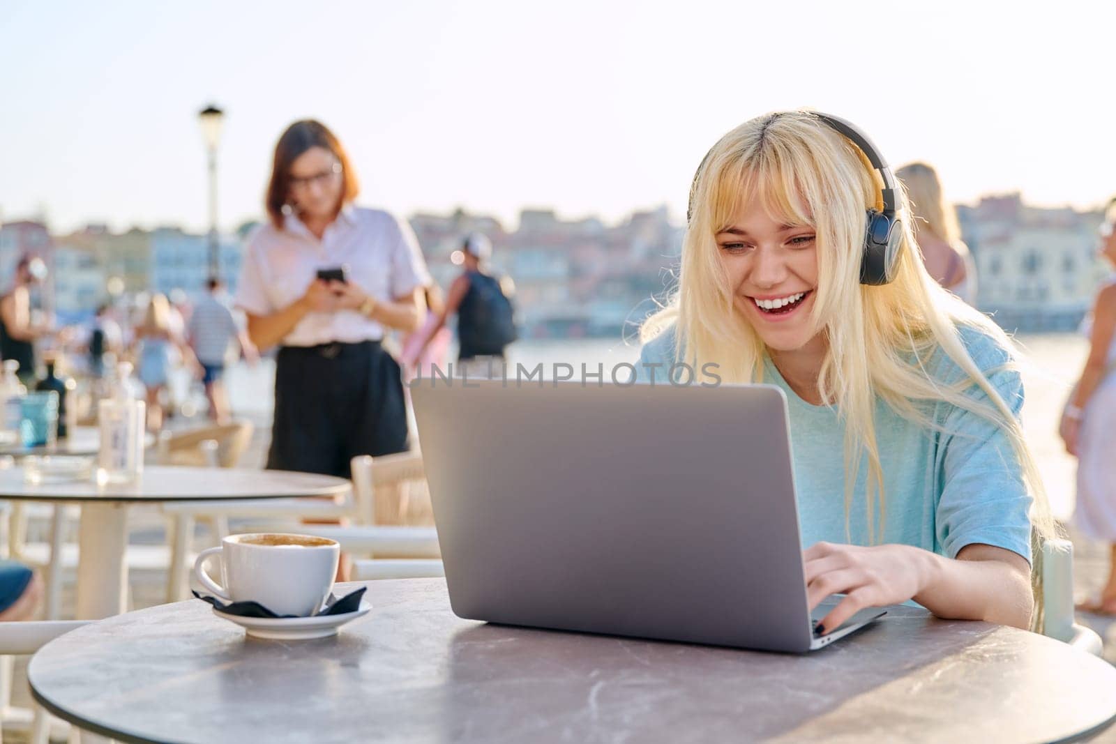 Smiling beautiful teenage girl in headphones looking into laptop. Blonde female in an outdoor cafe on waterfront using laptop. Youth, technology, lifestyle concept