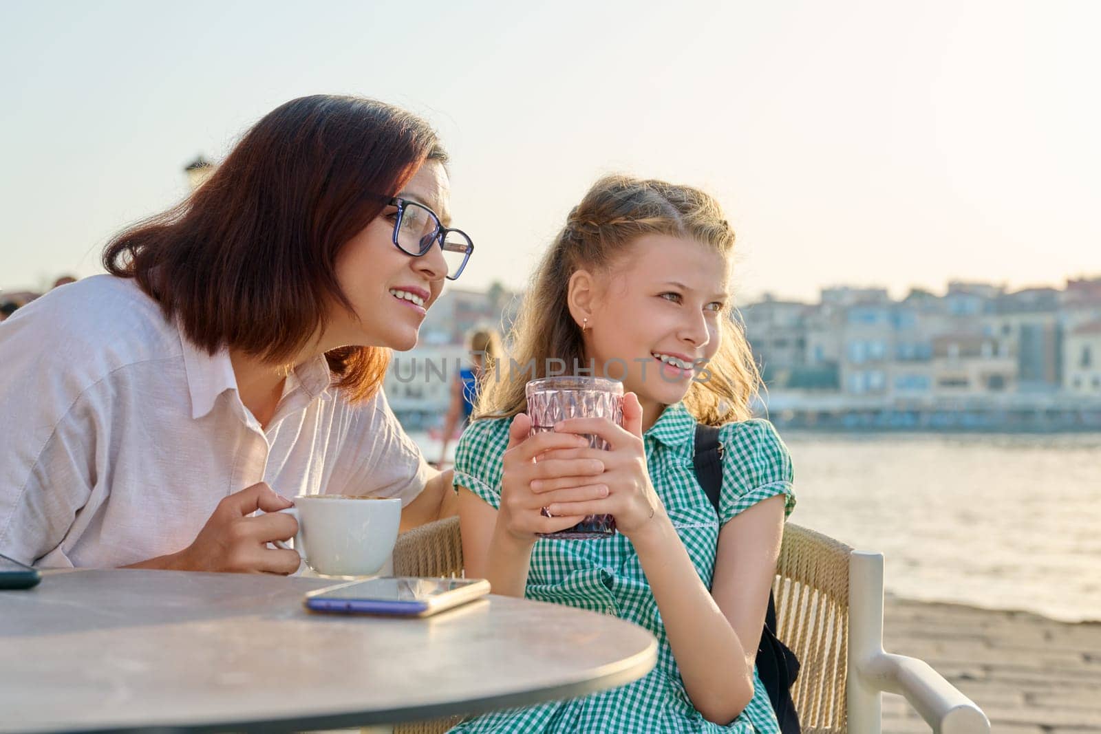 Happy mom and child daughter are resting together. Mother and preteen girl talking laughing in outdoor cafe, seaside city embankment background. Lifestyle, family, friendship, parent and kid concept