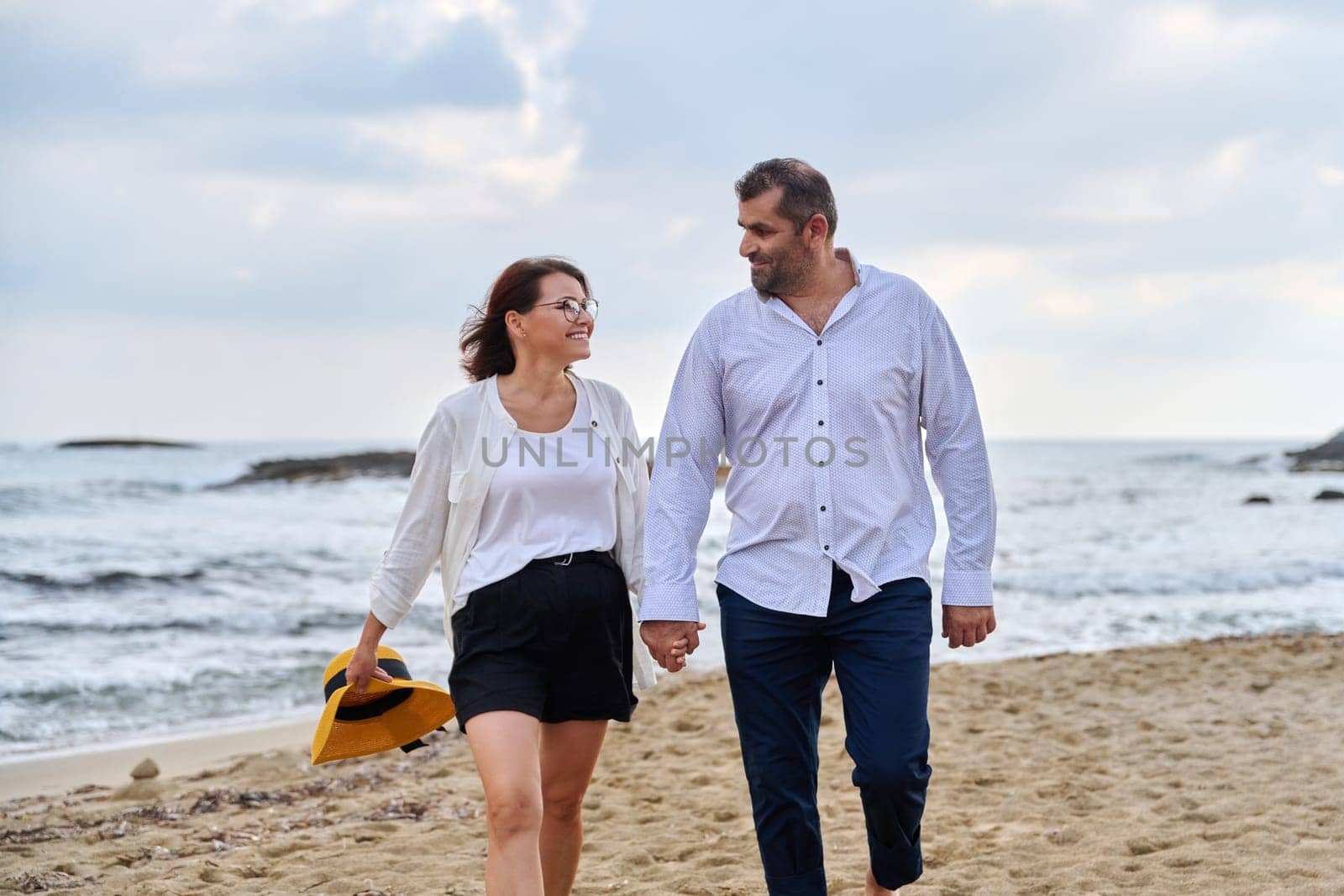 Happy middle-aged couple walking together on the beach. Mature man and woman holding hands walking along seashore. Relationship, lifestyle, vacation, tourism, sea nature, people concept