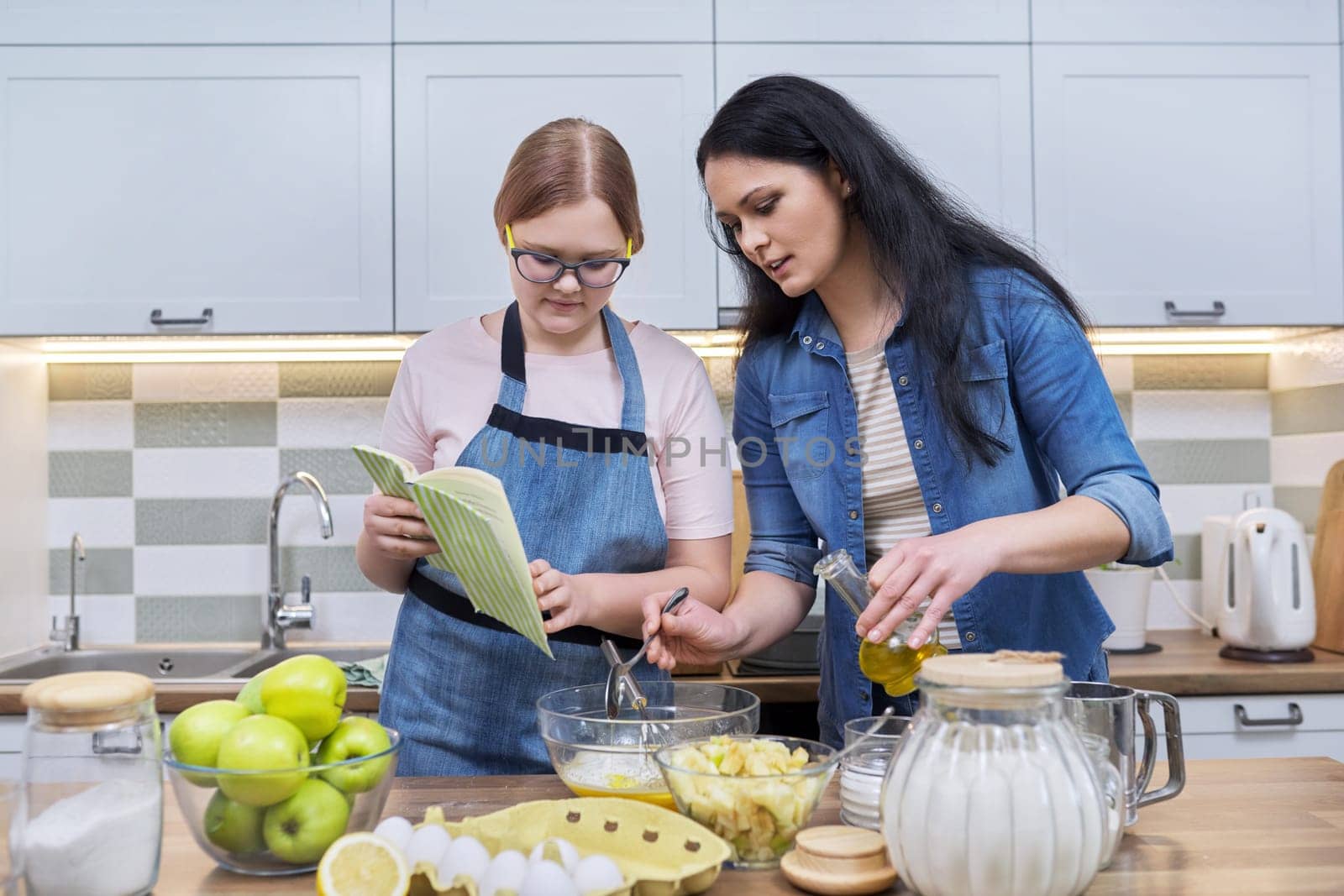 Mother and teenage daughter cooking at home in kitchen. Mom and girl making apple pie together, talking, use recipe book. Relationships, communication parent teenager, healthy homemade food, family