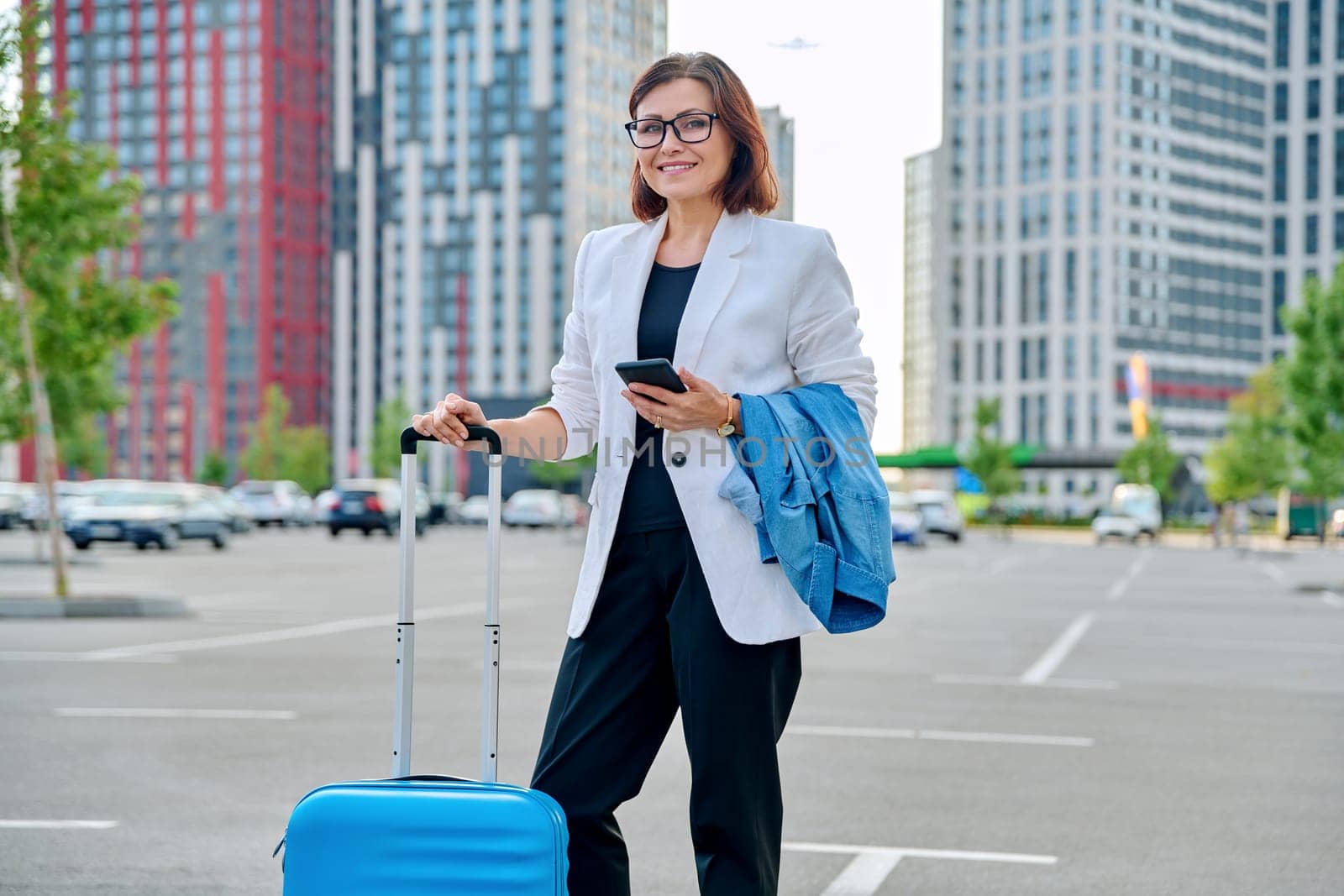 Middle aged confident business woman with suitcase, urban background. Smiling mature woman with smartphone luggage, looking at camera. Business trip, travel, city, work, lifestyle, people 40s concept