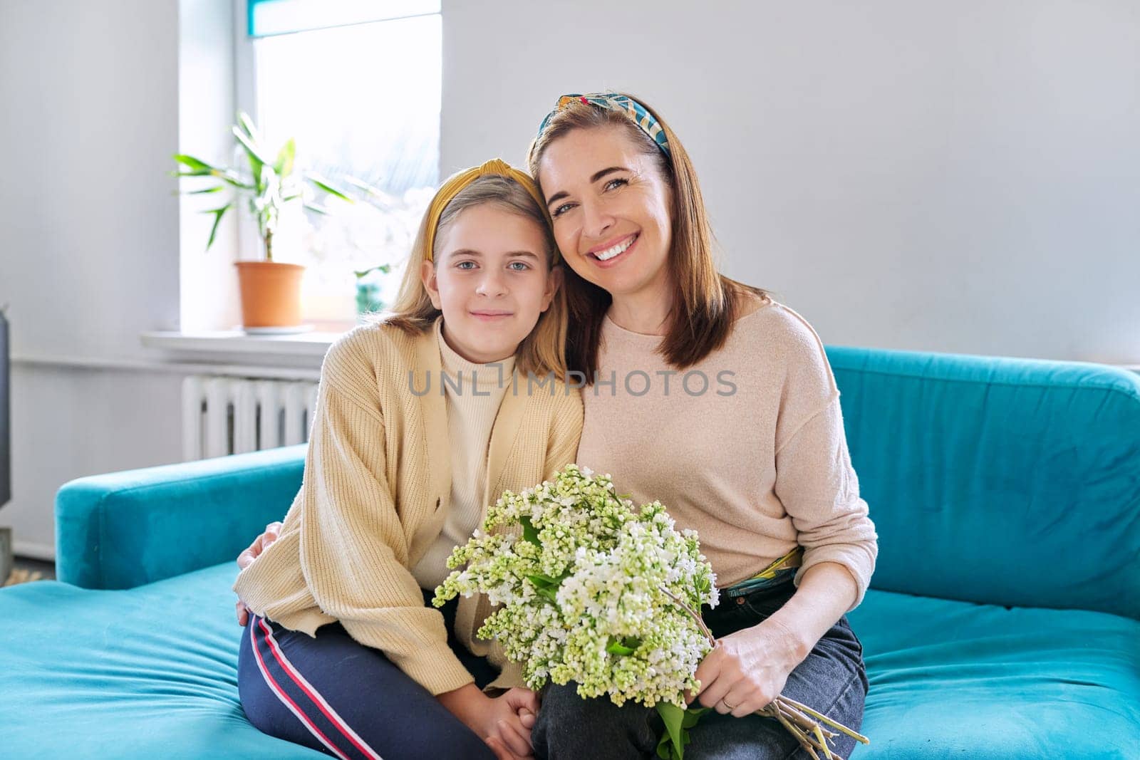 Happy mother and daughter child congratulating with bouquet of flowers and hand-drawn card, at home in living room on sofa. Mother's day, family, relationship, emotions, love, togetherness concept