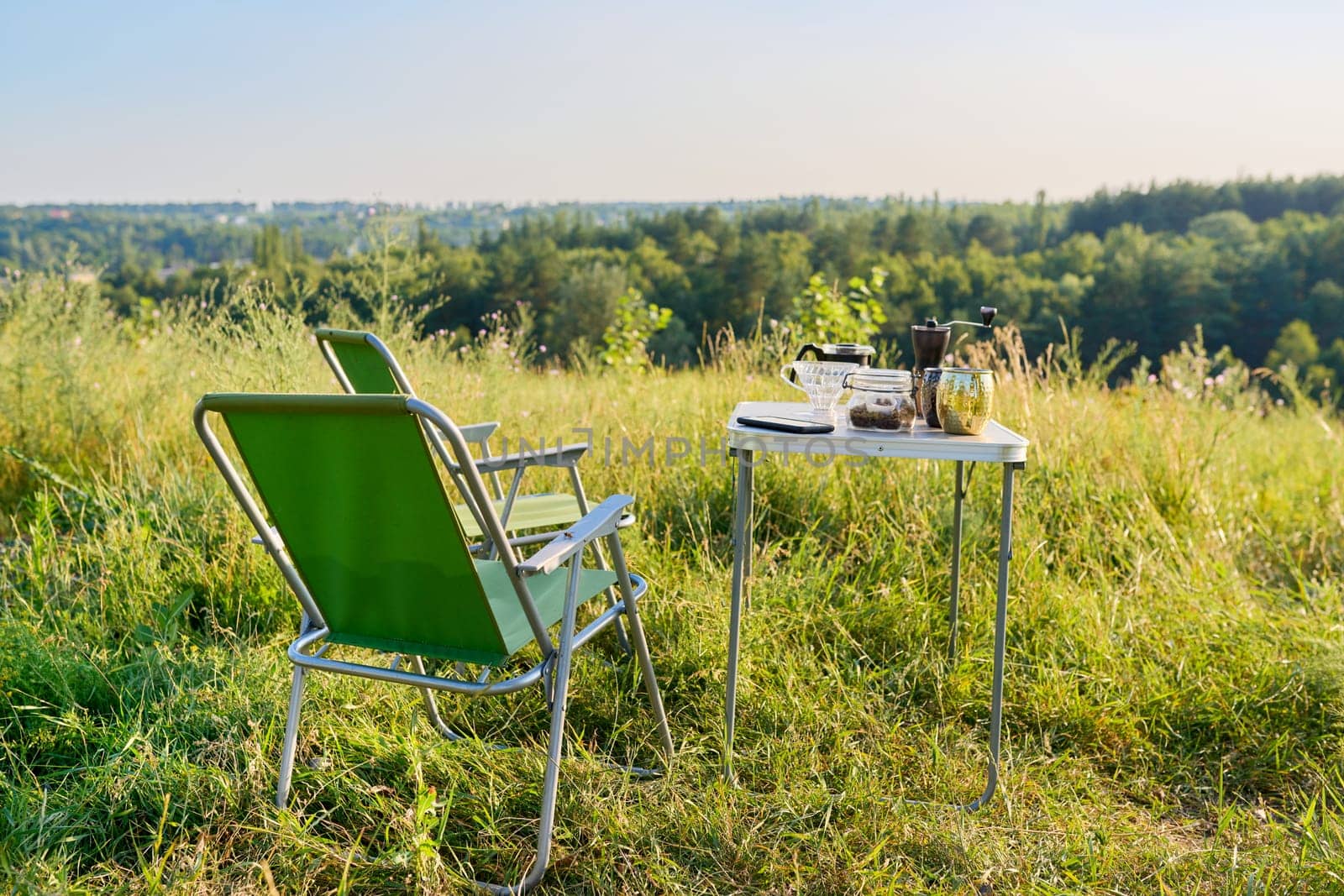 Set of folding furniture for camping, table and chair, cups utensils for brewing fresh coffee, summer nature of wild meadow, sunset on horizon. Summer, holidays, weekends, lifestyle, camping concept