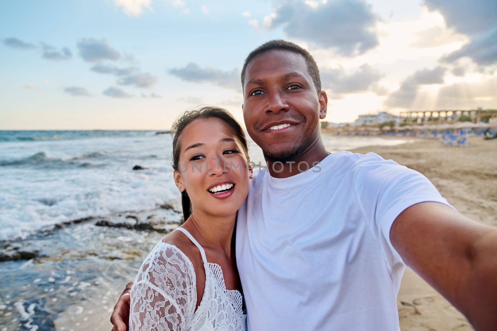 Happy couple in love taking selfie together on smartphone on beach, looking at camera. Vacation tourist trip to seaside resort, summer vacation, multicultural family, happiness, date, love concept