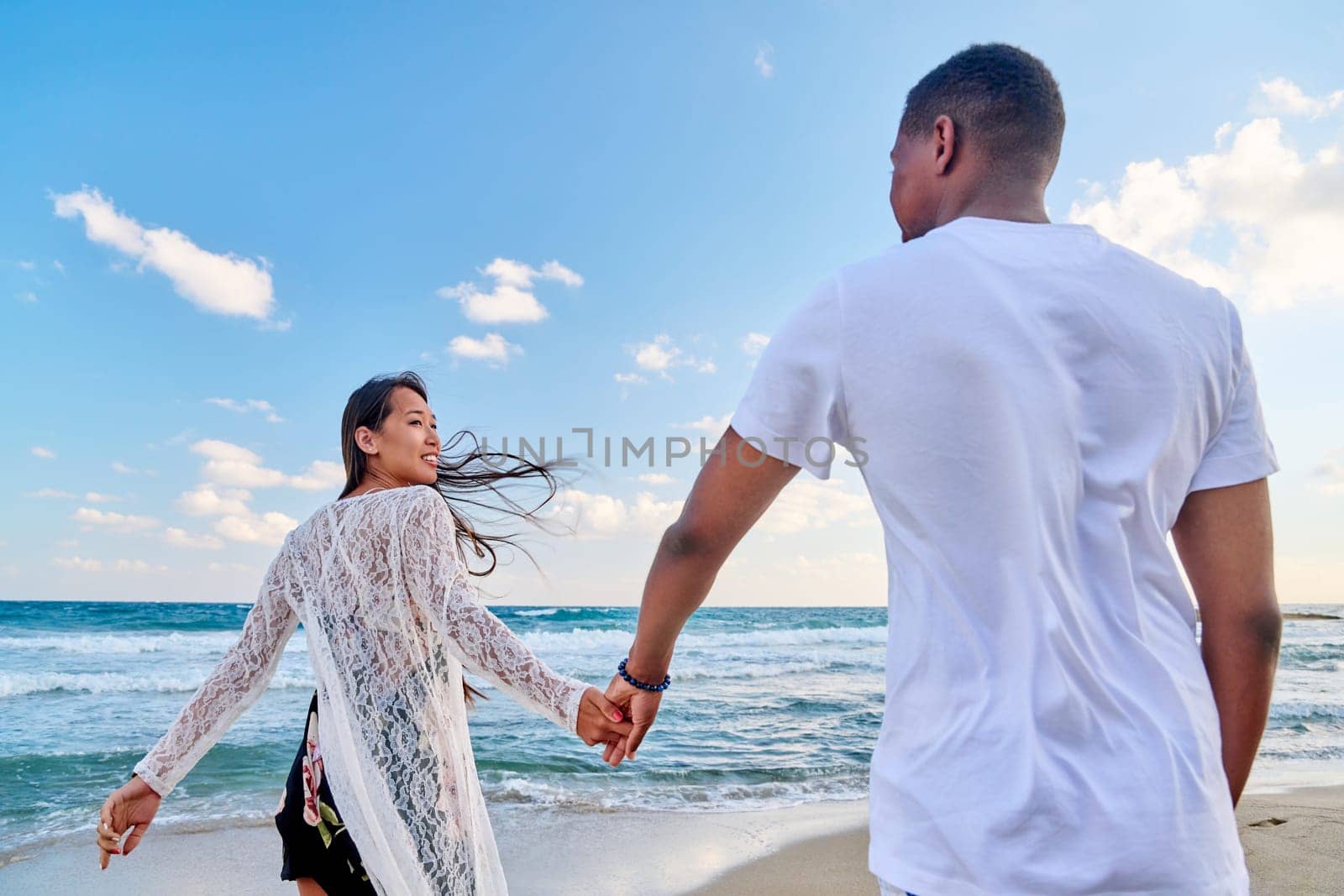 Loving happy couple walking holding hands on beach. Asian woman holding man's hand, sea sky background. Love, relationship, vacation, tourism, tourist travel to seaside resort, honeymoon concept