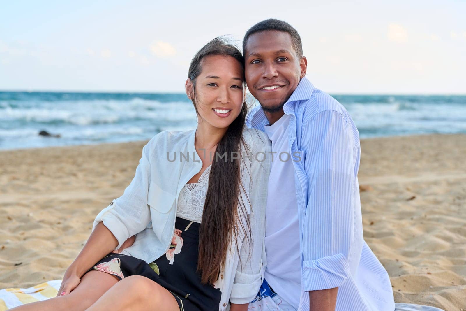 Happy multiethnic family resting on beach together. Asian woman and African man seated hugging each other on sand. Relationships, vacation travel and tourism at seaside resorts, people 20 30 years old