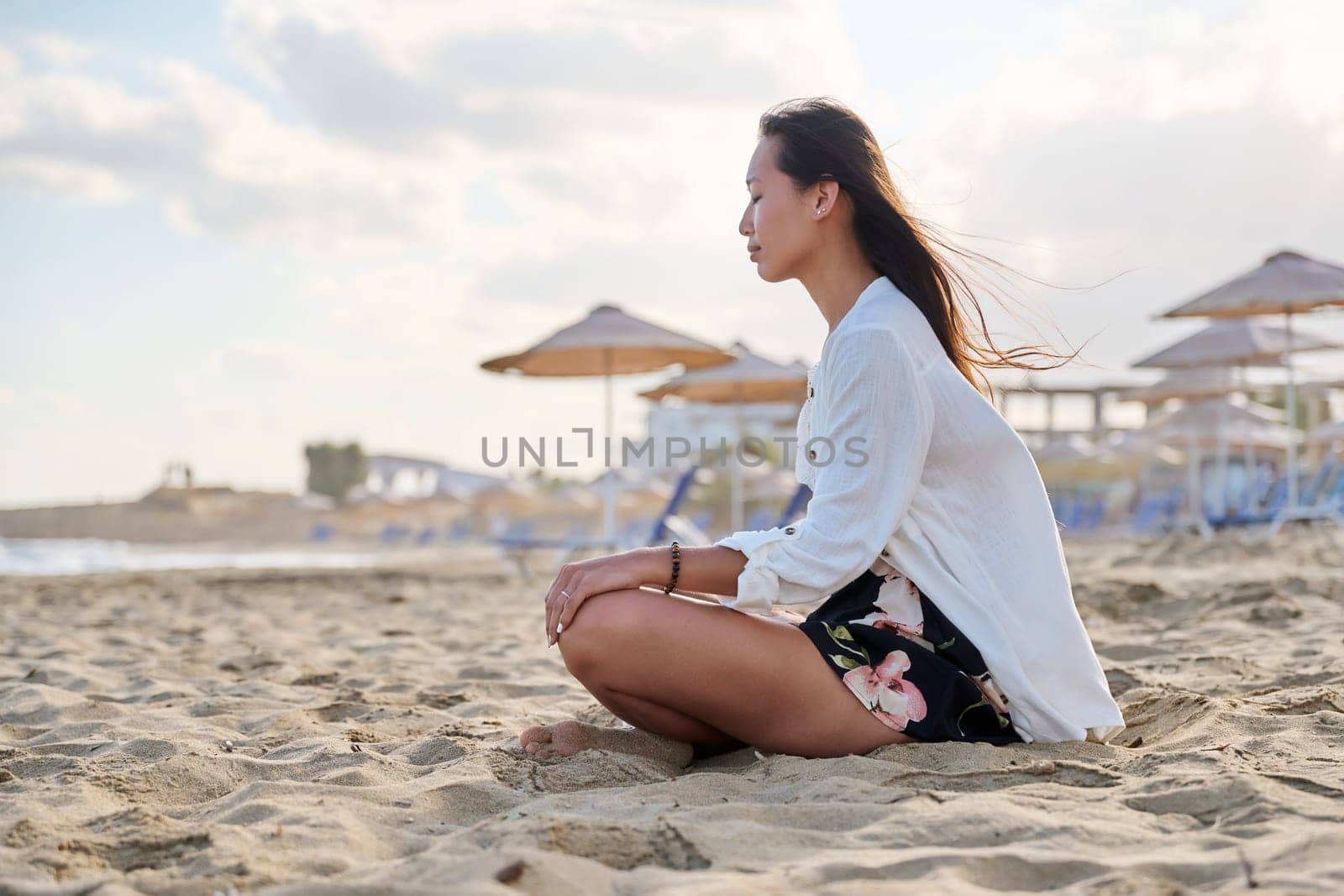 Young relaxed woman sitting in lotus position on beach, morning sea nature background. Natural Asian woman with closed eyes on sand. Meditation harmony, care for mental physical health, energy, people