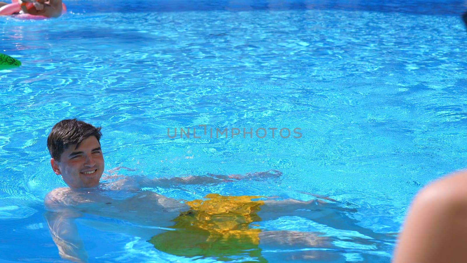 Young happy man swimming in the swimming pool in front of his girlfriend. Clip. Concept of travelling and vacation