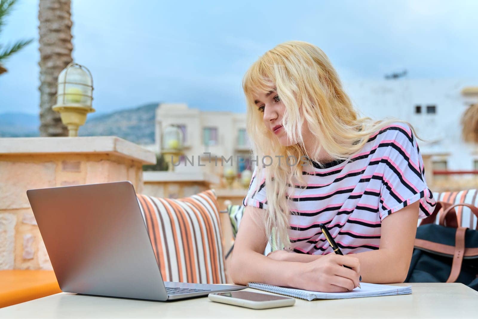 Teenage student sitting at outdoor table, using laptop, writing in notebook. Teenager female blonde with backpack, studying remotely, individually. Education knowledge technology, high school, college