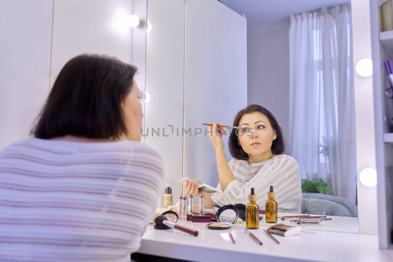 Beautiful middle aged woman doing makeup in front of a mirror. Cosmetics, beauty, make-up, face care, skin care, women 40s age concept