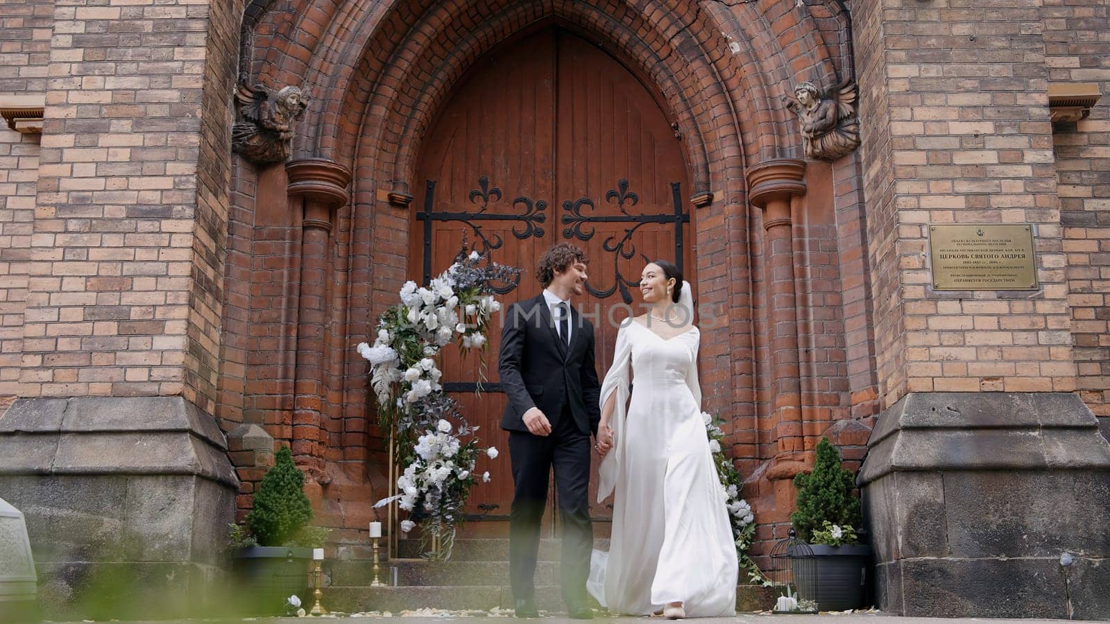 A bright and spectacular wedding couple. Action.Newlyweds where a bride in a white dress with a slit on her leg and a man with a beard in a suit walking next to a white arch with fresh flowers by Mediawhalestock