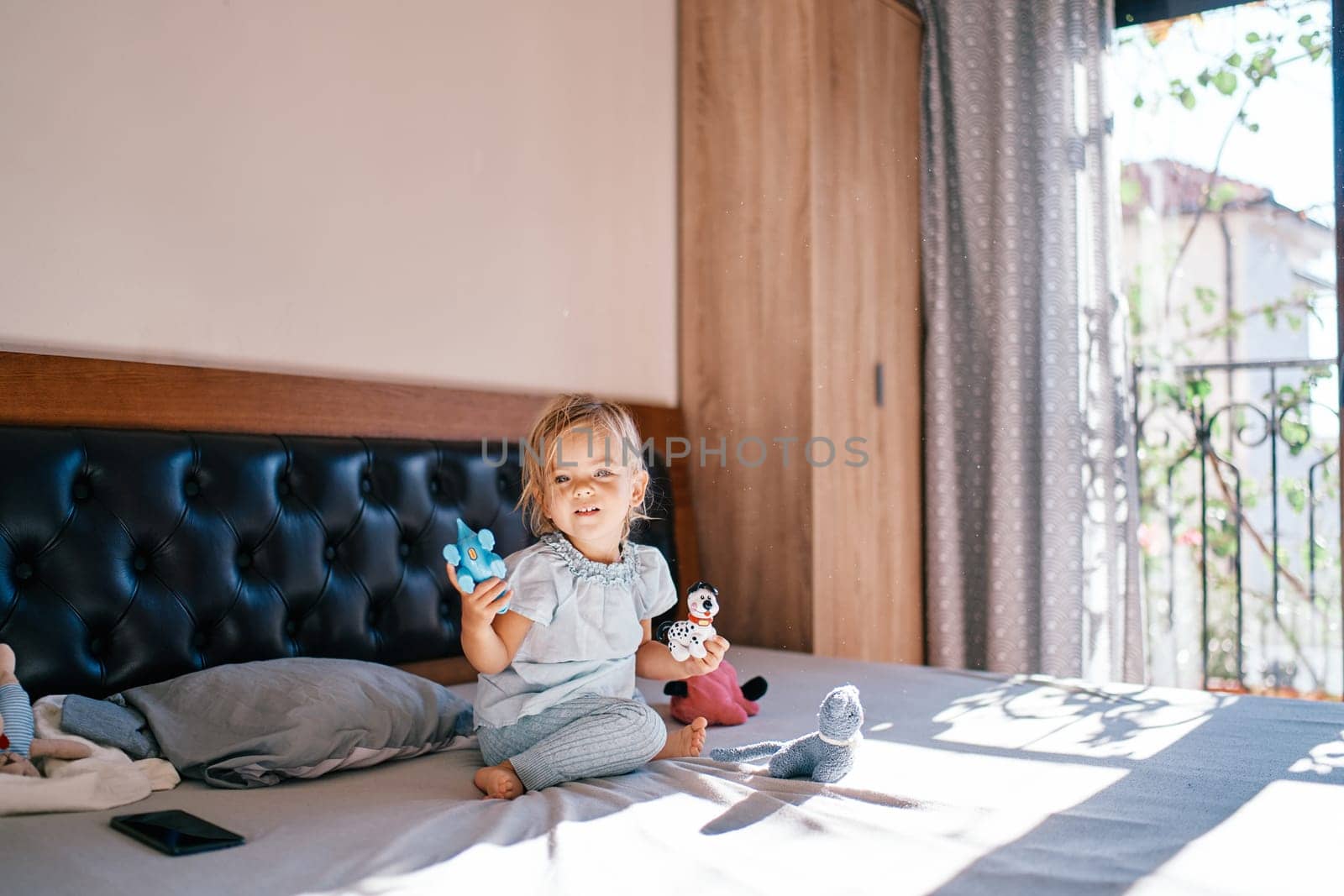 Little smiling girl sitting on the bed with toys in her hands. High quality photo
