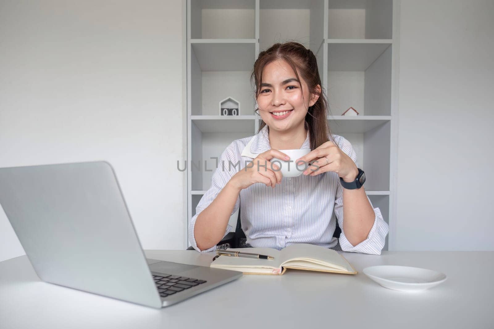 Young office worker sitting at table drinking coffee while checking documents and working on laptop..