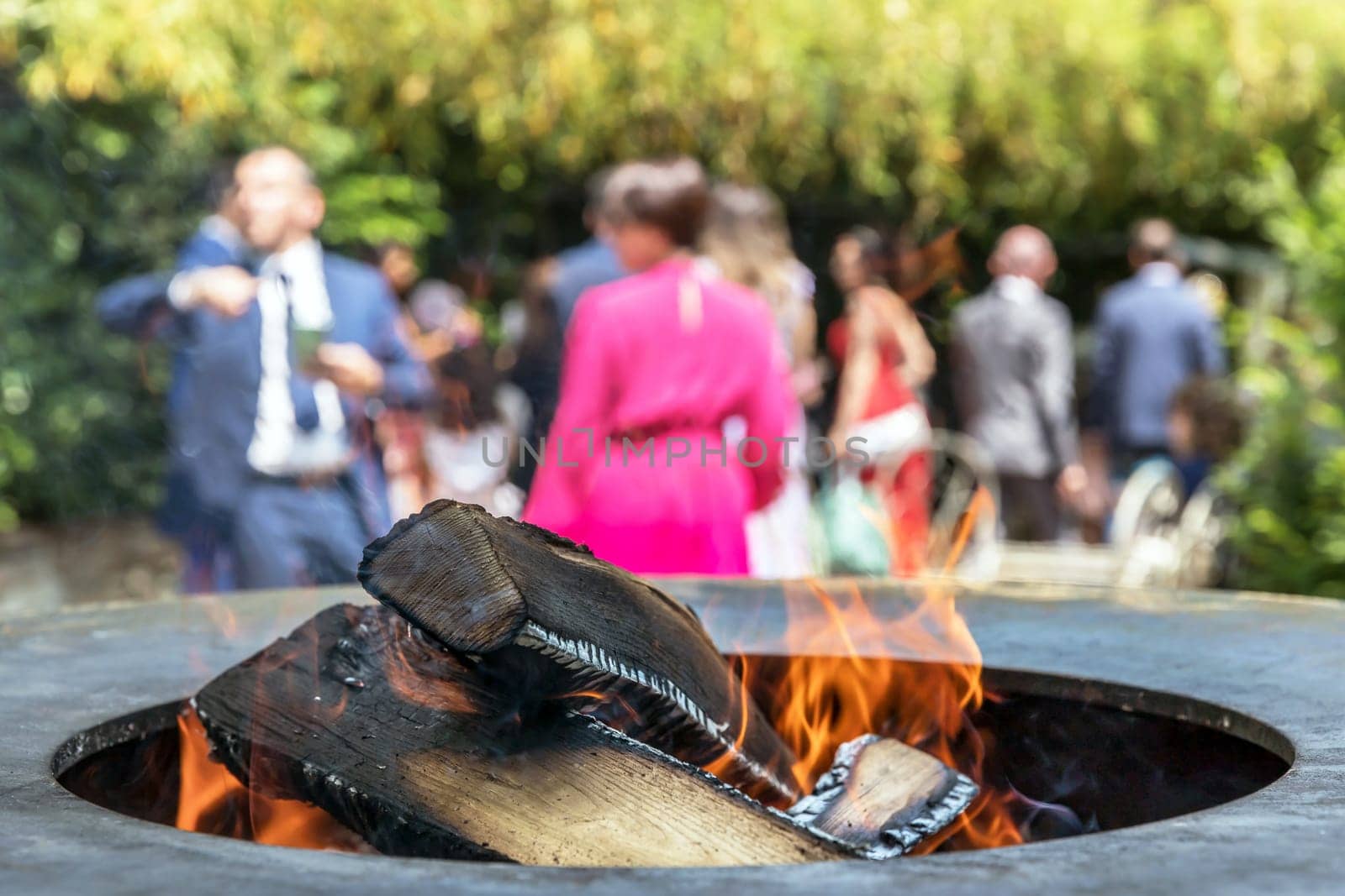 Close up shot of log pieces and fire wood, charcoal and ashes burning in flames in an old vintage brazier. In the blurred background party guests.
