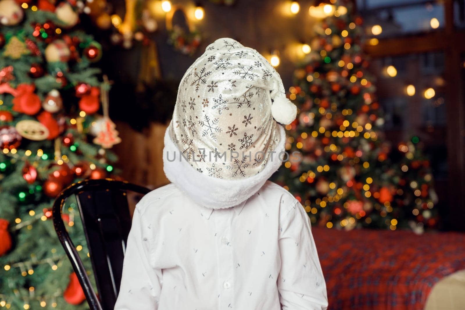 unrecognizable child in white shirt and santa hat stands on christmas tree background. Christmas celebration and fun.Faceless portrait. Boy with Christmas hat on his face. by YuliaYaspe1979