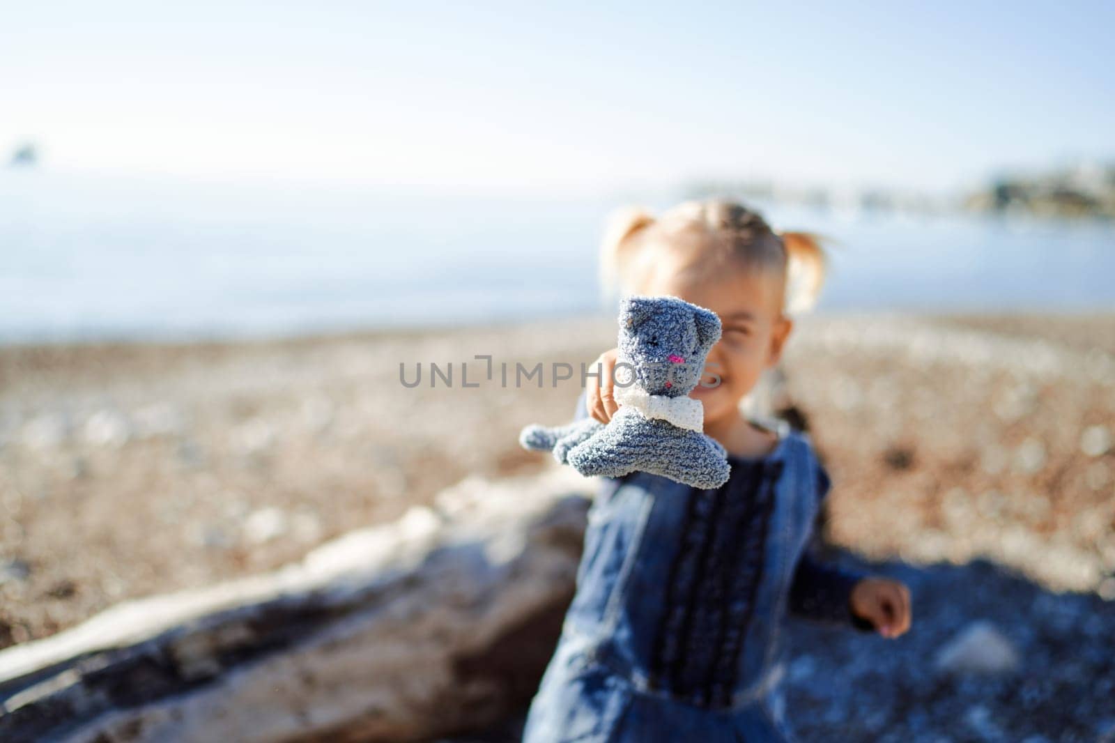 Little smiling girl stands near a driftwood on a sunny beach and holds out a toy by Nadtochiy