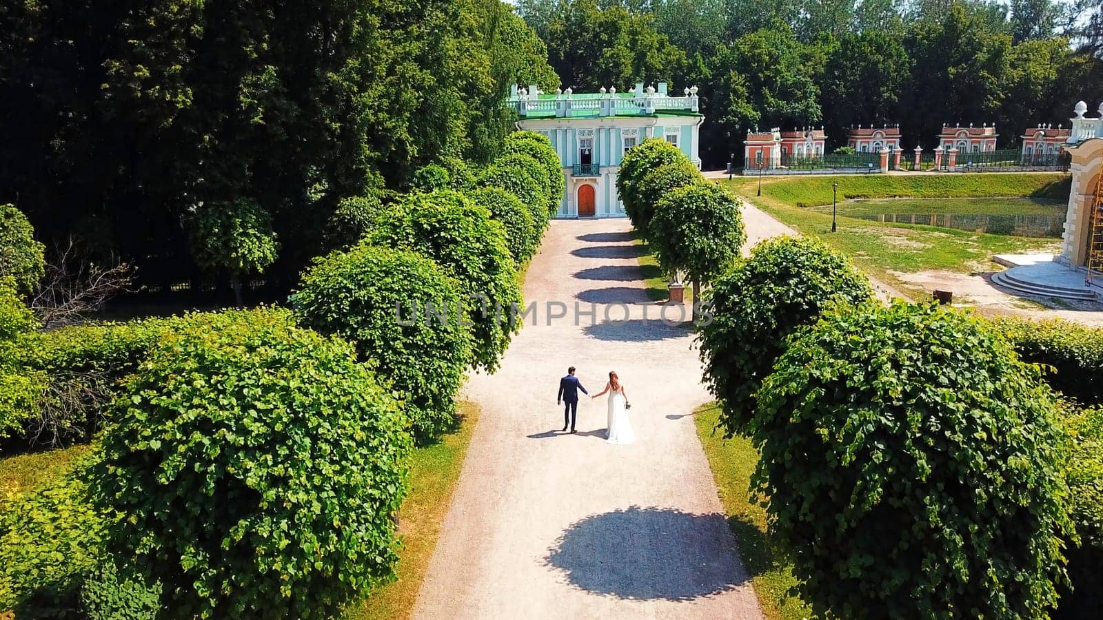 The view from the drone.Creative.A beautiful summer park with small beautifully trimmed trees and a summer road on which beautiful buildings and museums are visible and there is a young married couple with a bride in a beautiful dress and a husband in a suit. by Mediawhalestock