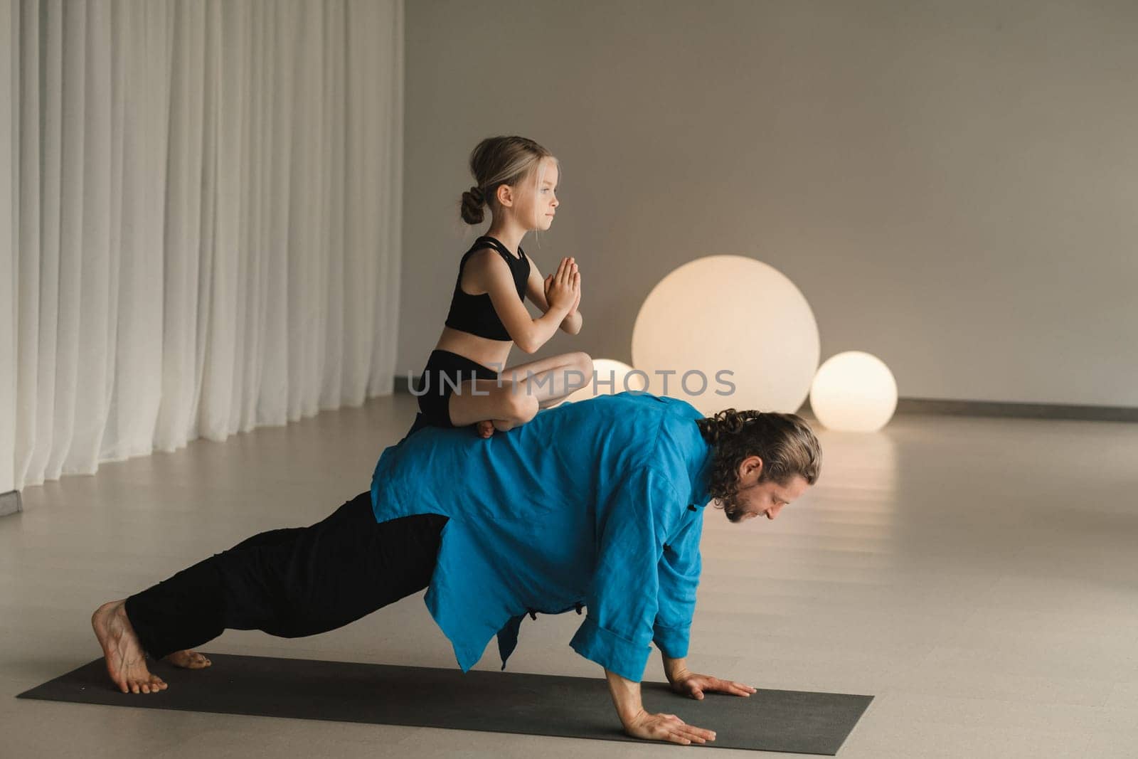 a child in the lotus position sits on the back of a yoga coach during training by Lobachad