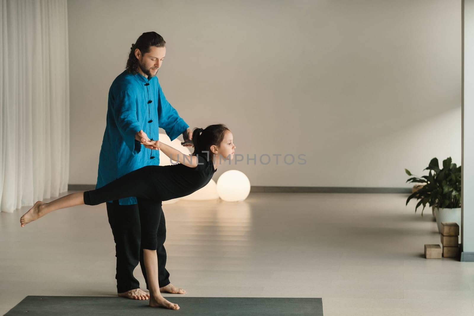 A yoga instructor in training helps a child to do exercises in the gym by Lobachad