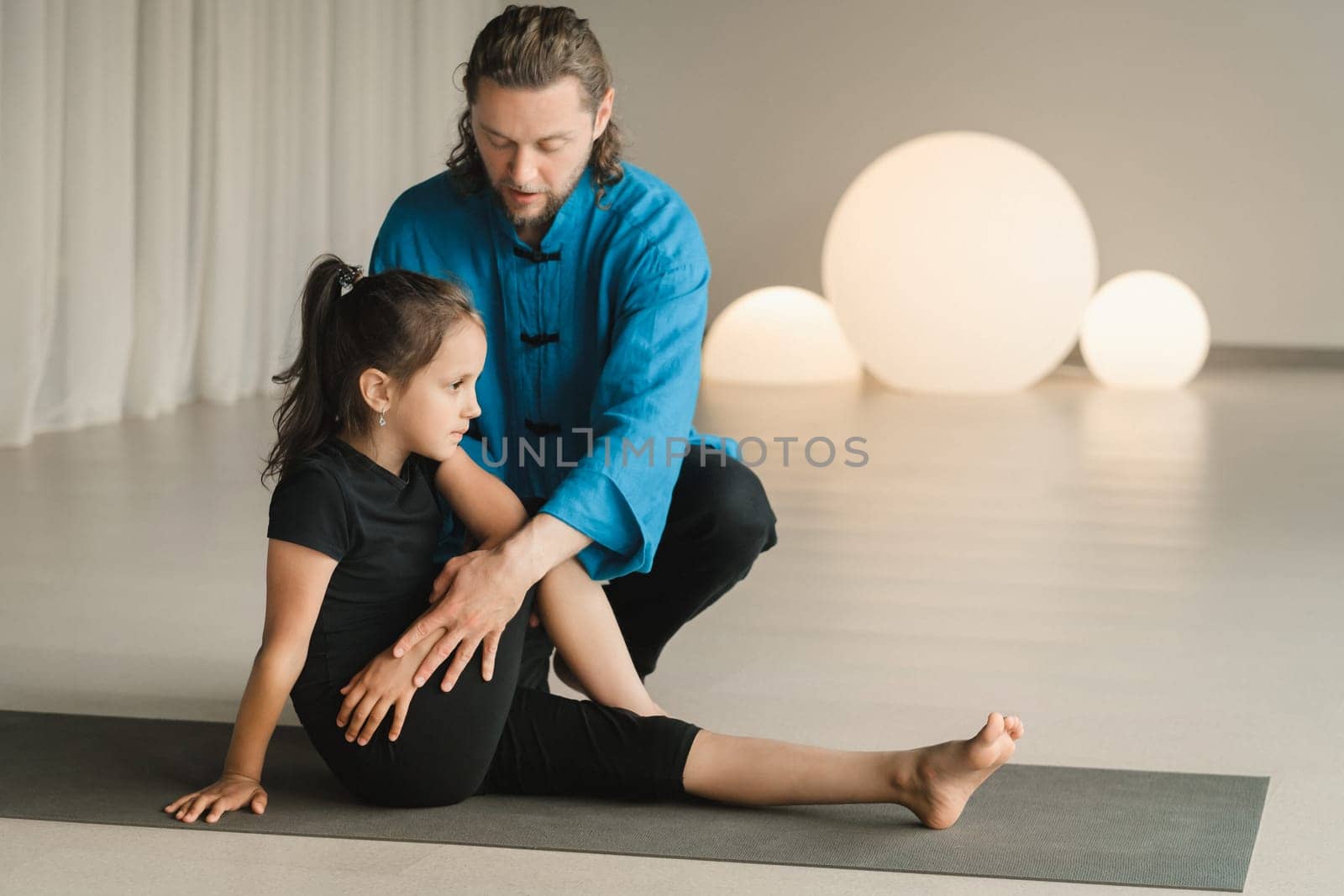 A yoga instructor in training helps a child to do exercises in the gym by Lobachad