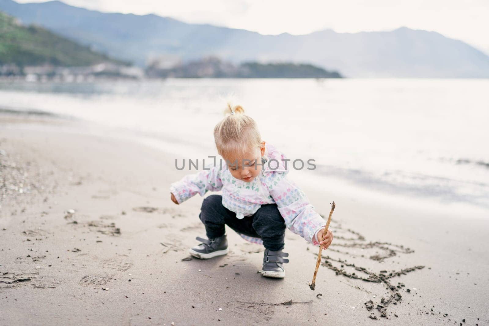 Little girl squats on the beach and draws with a stick on the sand. High quality photo
