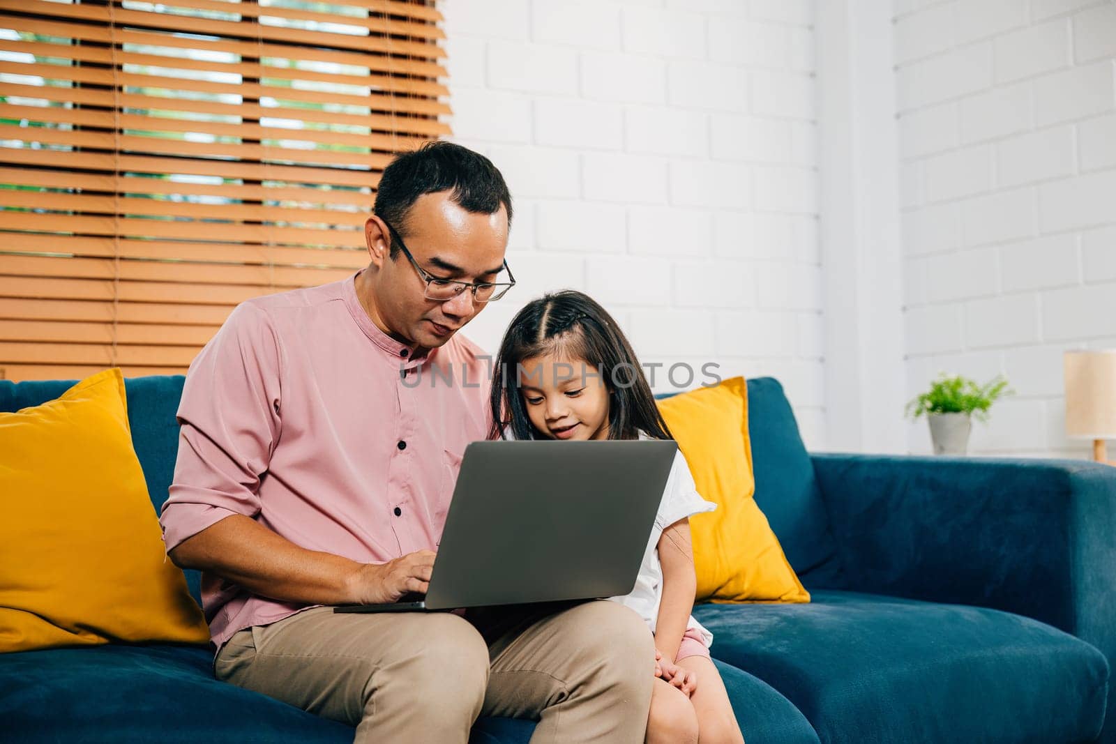 Smiling Asian father balances work and family with his daughter on laptop for e-learning creating special bonding moment on sofa. In their modern living room happiness and togetherness fill the air.