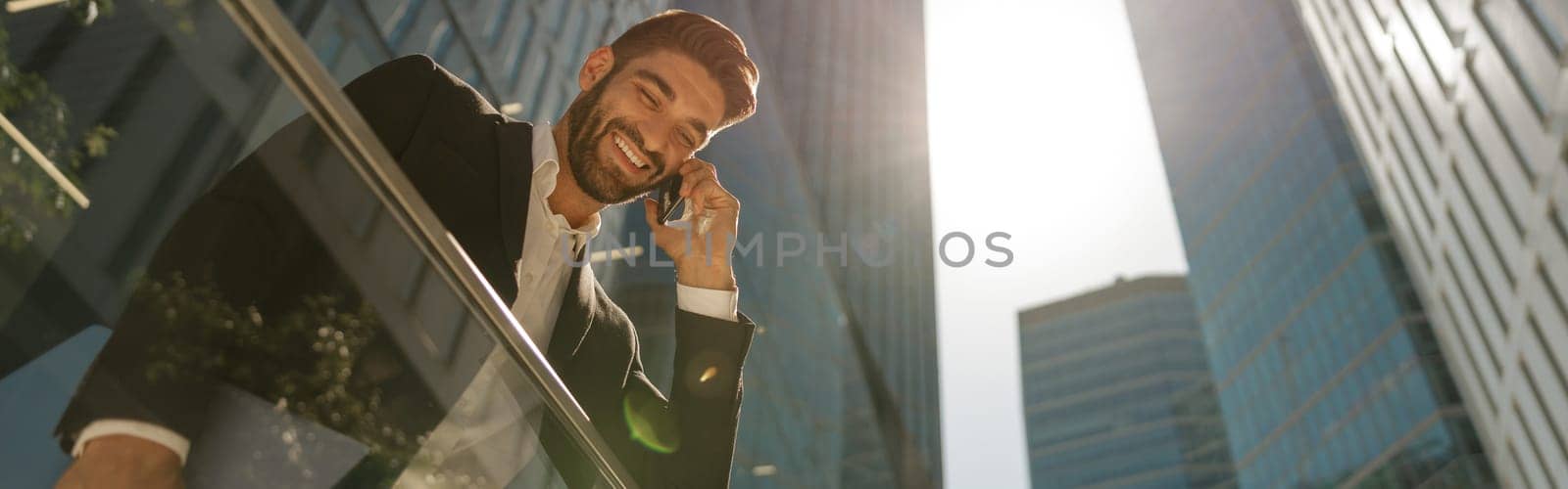 Bottom view of handsome entrepreneur using mobile phone standing on background of skyscrapers by Yaroslav_astakhov