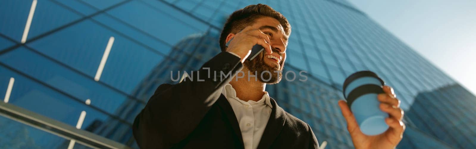 Handsome businessman is talking phone and drinking coffee standing on background of city skyscrapers by Yaroslav_astakhov