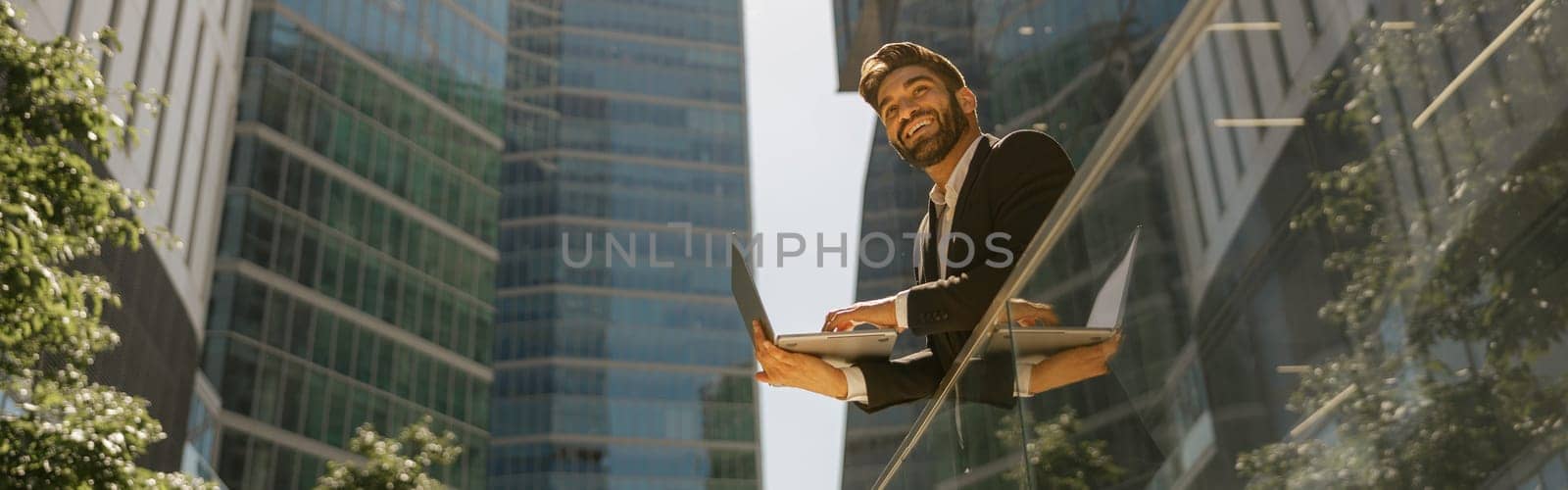 Stylish male manager working on laptop standig on office terrace and looks away. High quality photo