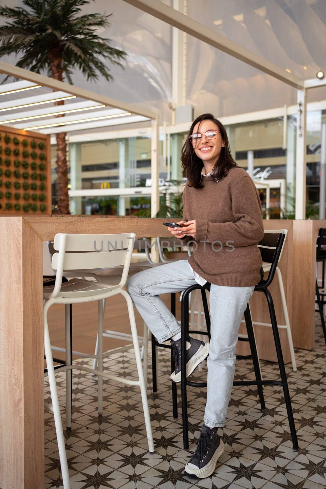 Woman sits in modern cafe during lunch wearing glasses look in camera. Female employee came in for snack in cafeteria at his office. Business woman smiles and sits in cafe with phone waiting