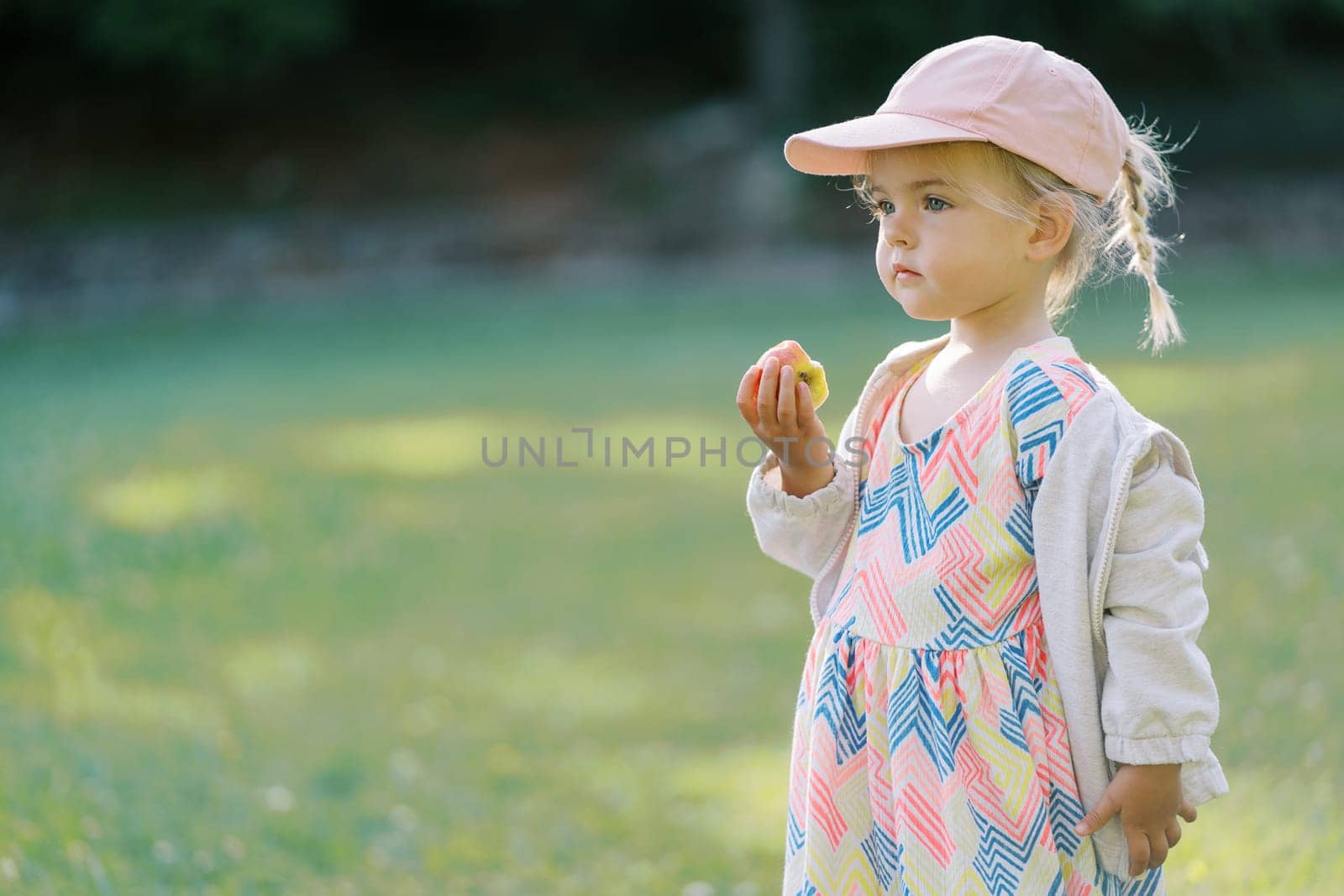 Little girl with a bitten apple in her hand stands in a clearing. Side view by Nadtochiy